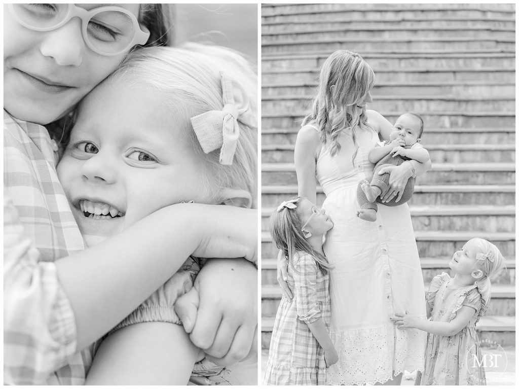 Black and white image of two sisters hugging tightly and looking up at mom holding baby brother on Airlie steps at their Fall Mini Session taken by TuBelle Photography, a DMV Photographer.