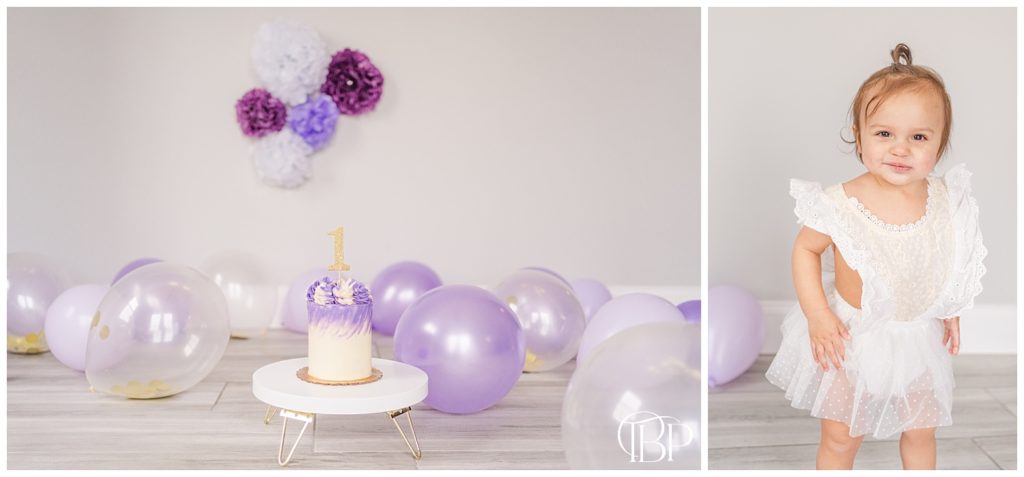 Little girl in beige, lace onesie and cake surrounded by purple and white balloons for cake smash session. Taken by TuBelle Photography, a Haymarket, Virginia cake smash photographer.