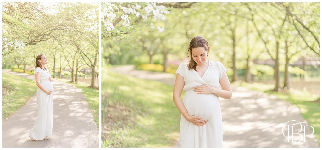 Expecting mom in white dress holds her baby bump on a park path for a maternity session at Meadowlark Botanical Gardens in Fairfax County, VA taken by TuBelle Photography, a Vienna, Virginia maternity photographer.