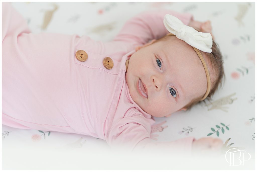 Newborn baby girl in pink onesie and white bow smiles at camera in her crib for her lifestyle newborn photos in Centerville, Virginia taken by TuBelle Photography.