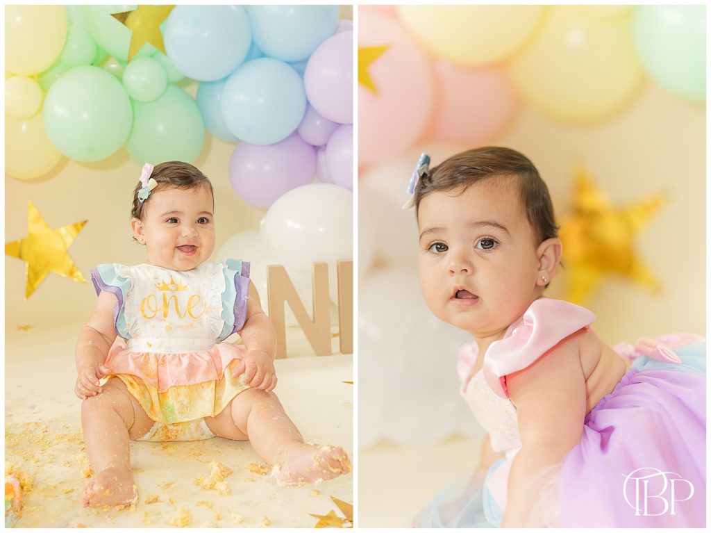 Baby girl smiling while sitting in cake crumbs for her rainbow themed cake smash pictures. Taken by TuBelle Photography, a Fairfax County, Virginia cake smash photographer.