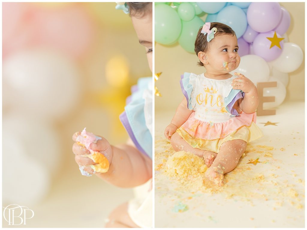 Baby girl squeezing cake and icing in her hand for her rainbow themed cake smash pictures in Fairfax County, VA. Taken by TuBelle Photography, a cake smash photographer.